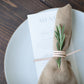 Handcrafted veg-tan Leather Napkin Tie by Keyaiira shown on a white ceramic plate with a tan linen napkin and fresh greenery, artisan-made leather cord for elegant, modern table styling.