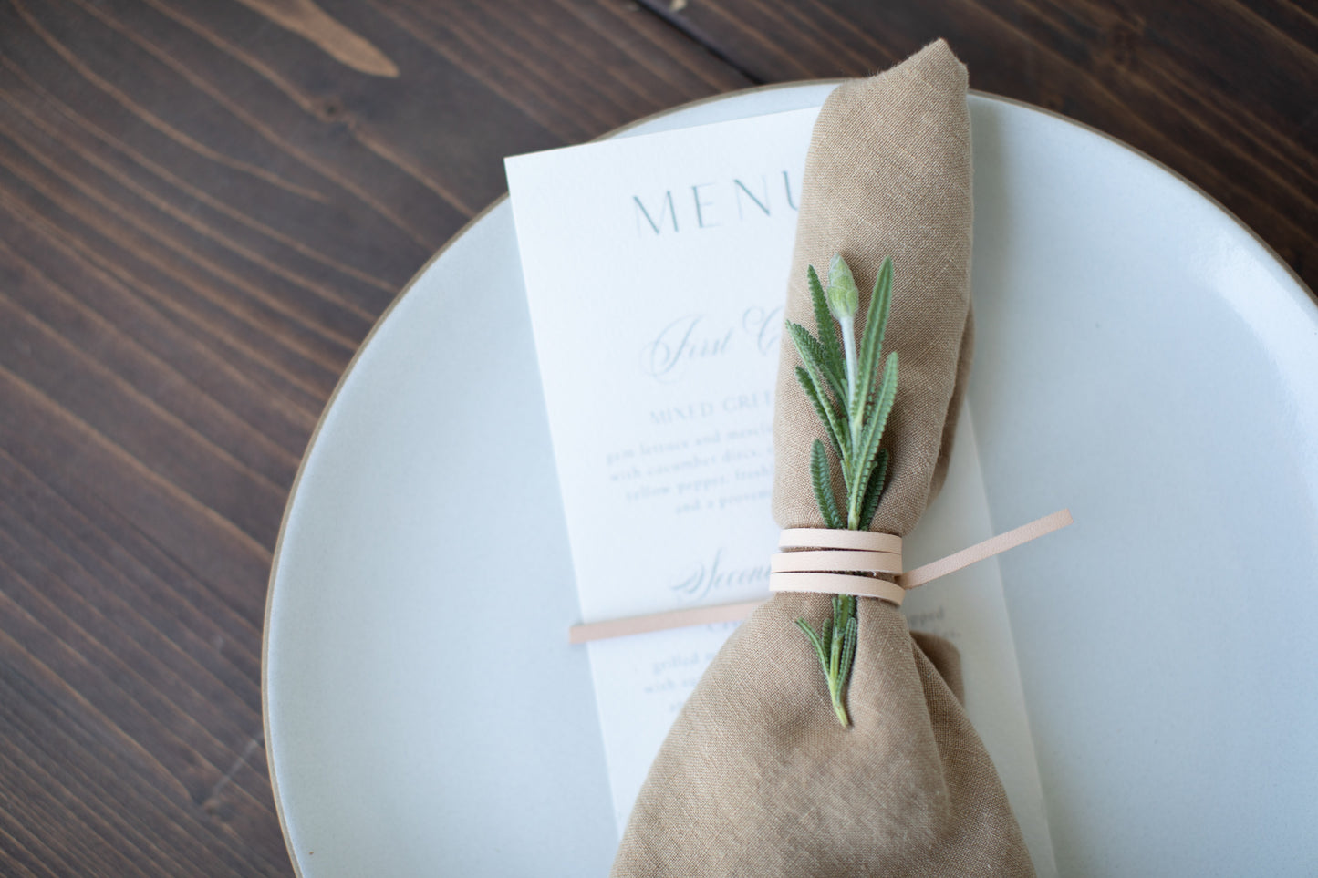 Handcrafted veg-tan Leather Napkin Tie by Keyaiira shown on a white ceramic plate with a tan linen napkin and fresh greenery, artisan-made leather cord for elegant, modern table styling.