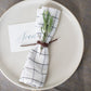 Artisan-made Leather Napkin Tie by Keyaiira in walnut leather cord paired with a white plaid napkin and greenery sprig for a warm, organic table presentation.