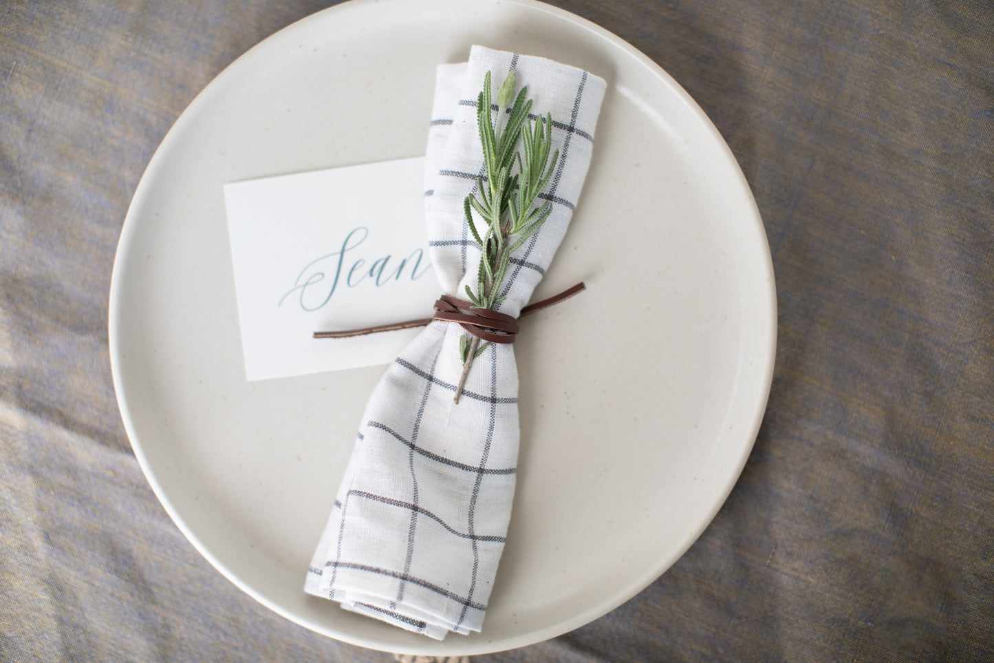 Artisan-made Leather Napkin Tie by Keyaiira in walnut leather cord paired with a white plaid napkin and greenery sprig for a warm, organic table presentation.