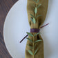 Wine-toned Leather Napkin Tie from Keyaiira. Soft, flexible leather cord wrapped around an earthy green napkin with eucalyptus sprig, adding a warm, modern touch to elegant dining decor.