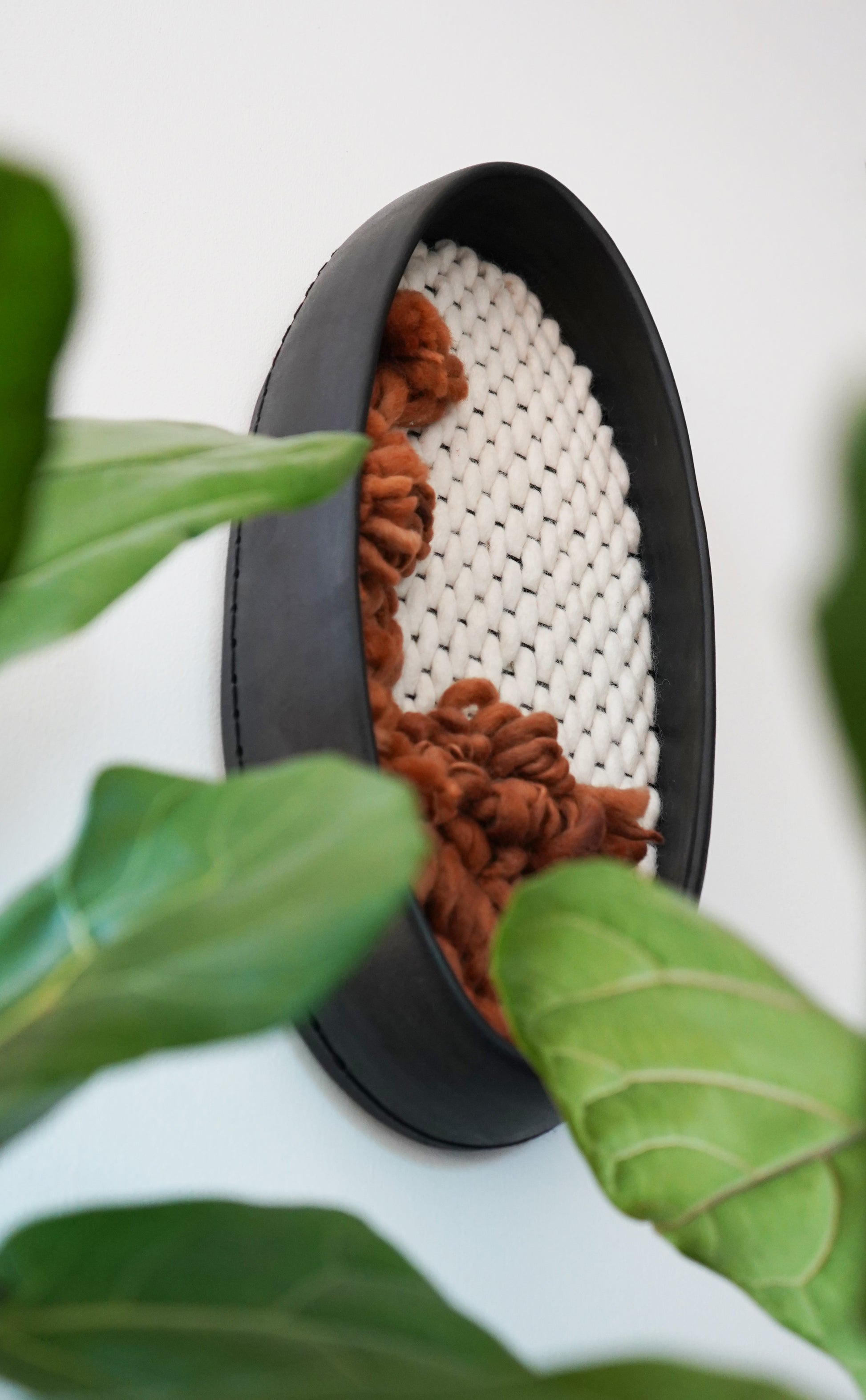 Modern organic wall decor hanging on a white wall, styled with greenery. The dark black frame and rust-toned wool add warmth and an earthy, grounding element to a minimalist or Japandi-style interior.