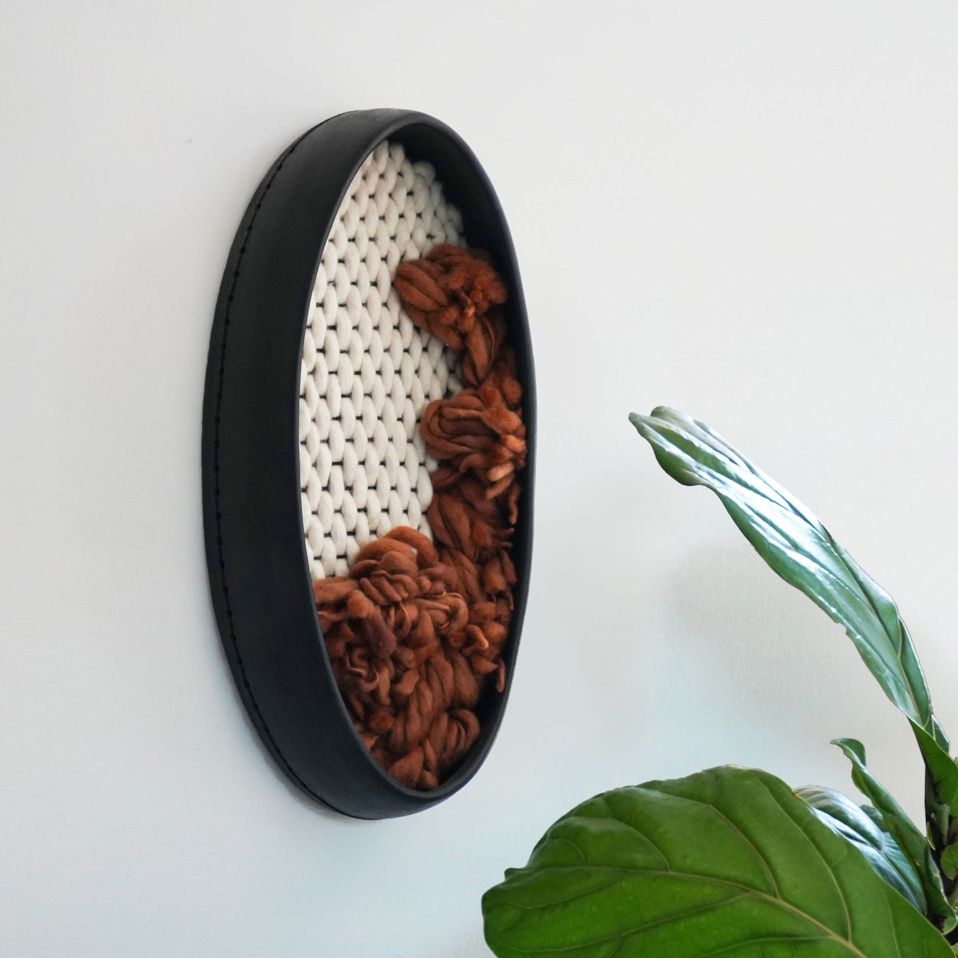 Handcrafted matte black ceramic clay frame wall sculpture sized 19x9.5 inches. The interior features a woven ivory wool grid with organic, rust-brown thick and thin wool yarn cascading from the center. Modern bohemian fiber art by Keyaiira.
