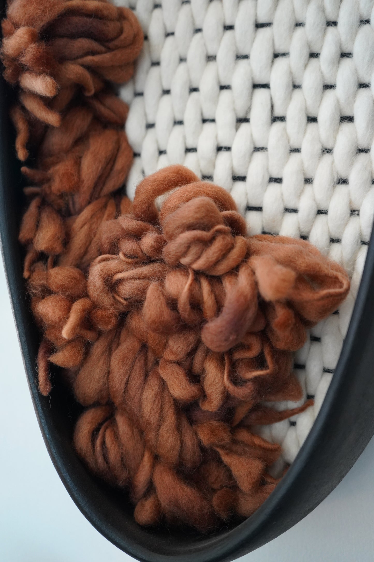 Macro detail of Woven Wall Hanging -  hand-dyed rust brown wool fibers contrasting against a soft white hand-felted wool background. Highlights the tactile texture of the New Zealand sheep’s wool and the matte finish of the black clay rim.