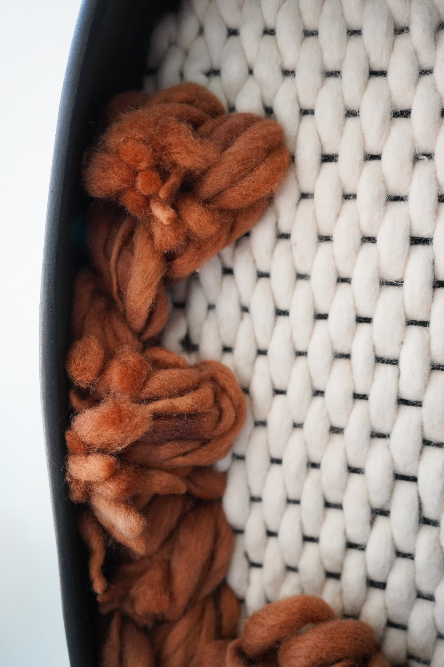Macro detail of Zenia wall sculpture texture. Hand-spun rust thick-and-thin wool yarn loops emerge from a hand-felted ivory New Zealand wool grid, framed by a sleek matte black ceramic clay rim.