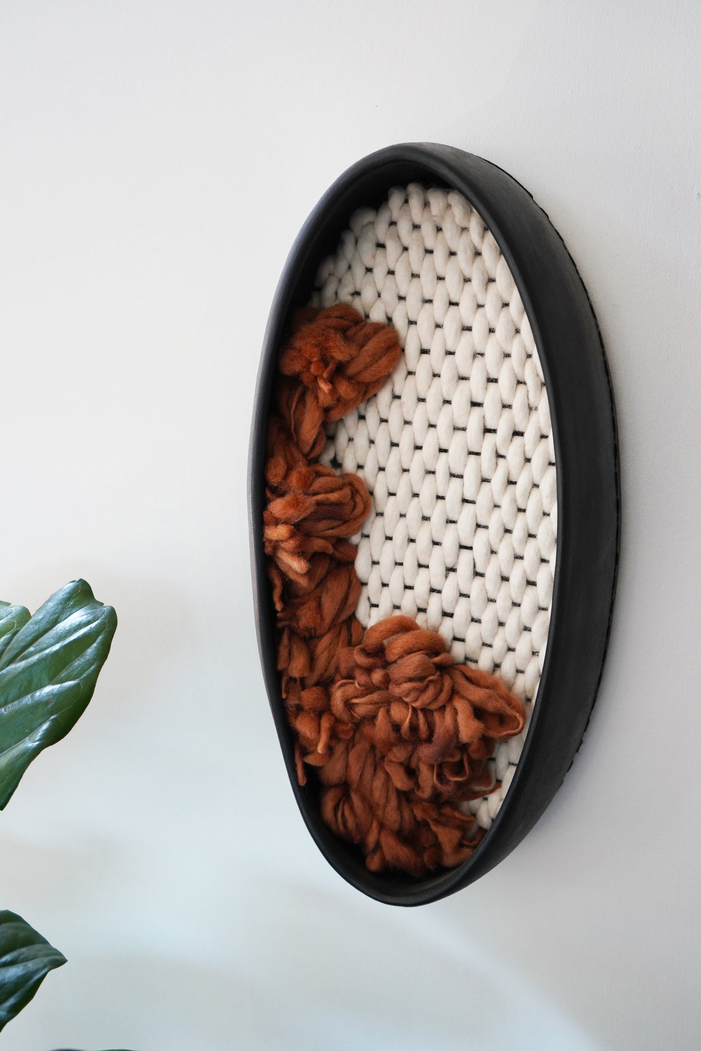 Side angle of black clay wall hanging showing the 3.25-inch depth of the ceramic frame. Demonstrates the dimensional quality of the fiber art where the rust wool emerges from the flat ivory woven base
