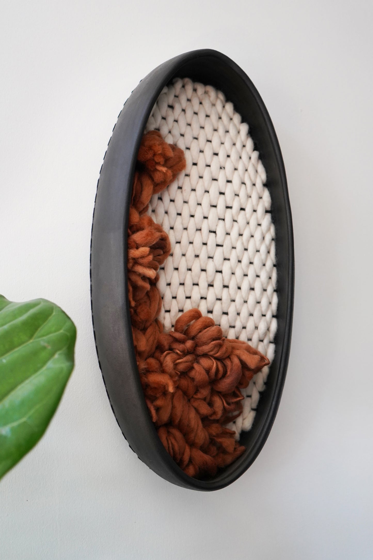 Angled view of oval ceramic wall art, capturing the hand-formed irregular shape of the black clay frame made in California. Shows the juxtaposition of hard ceramic and soft, hand-spun wool textures.