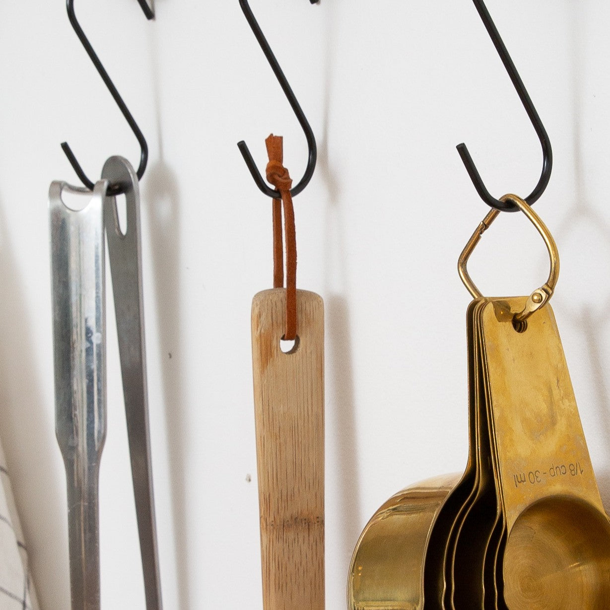 Detachable leather hanging strings by Keyaiira used to hang wooden and metal kitchen utensils on wall hooks — handcrafted leather hanging strings for functional home organization with a white wall behind it.