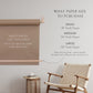Keyaiira kraft paper holder size guide displayed on a wall, showing recommended paper widths 18-inch small, 24-inch medium, and 30-inch large. Tan leather straps and birch dowel hold a roll of kraft paper above a woven chair, with text noting that kraft paper is not included and a securing strap prevents the paper from rolling up. Modern minimalist décor for home, office, or studio organization.
