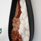 Angled view of the full sculptural piece showing the curvature of the terracotta clay frame and the combination of soft white wool, warm brown yarn, and avocado dyed fibers. Textural fiber and clay art for modern interiors.