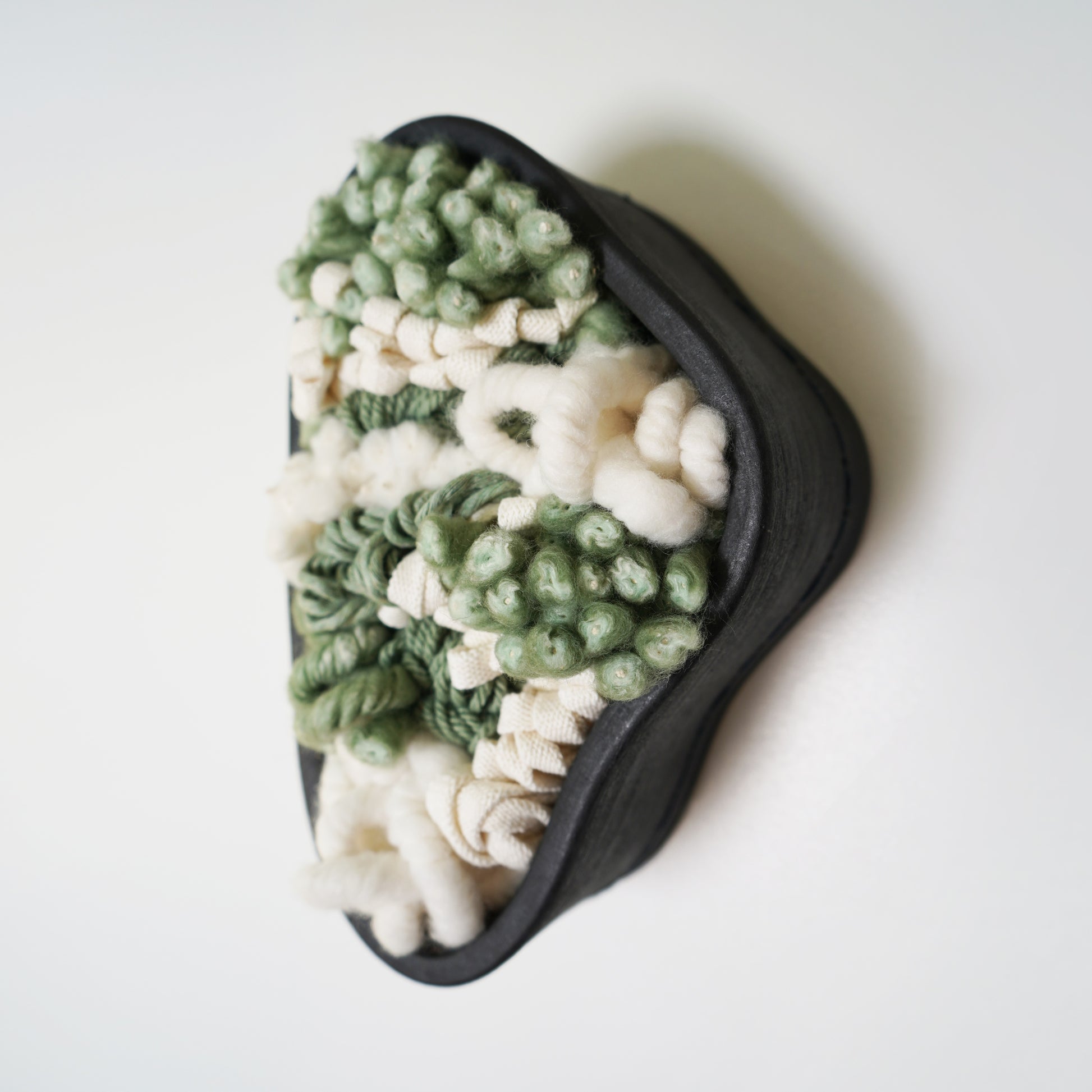 Full view of a handcrafted sculptural wall hanging featuring a brushed matte black clay frame. Soft, intricate fibers made from recycled plastic bottles and wool extend slightly beyond the ceramic edge, contrasting with the rigid frame.
