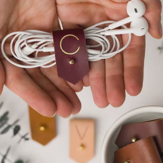 Keyaiira leather earbud holder demonstration showing a Wine C initial design and brass stud-and-post fastening for managing headphones, charger cords and everyday small cables
