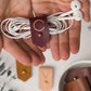 Keyaiira leather earbud holder demonstration showing a Wine C initial design and brass stud-and-post fastening for managing headphones, charger cords and everyday small cables
