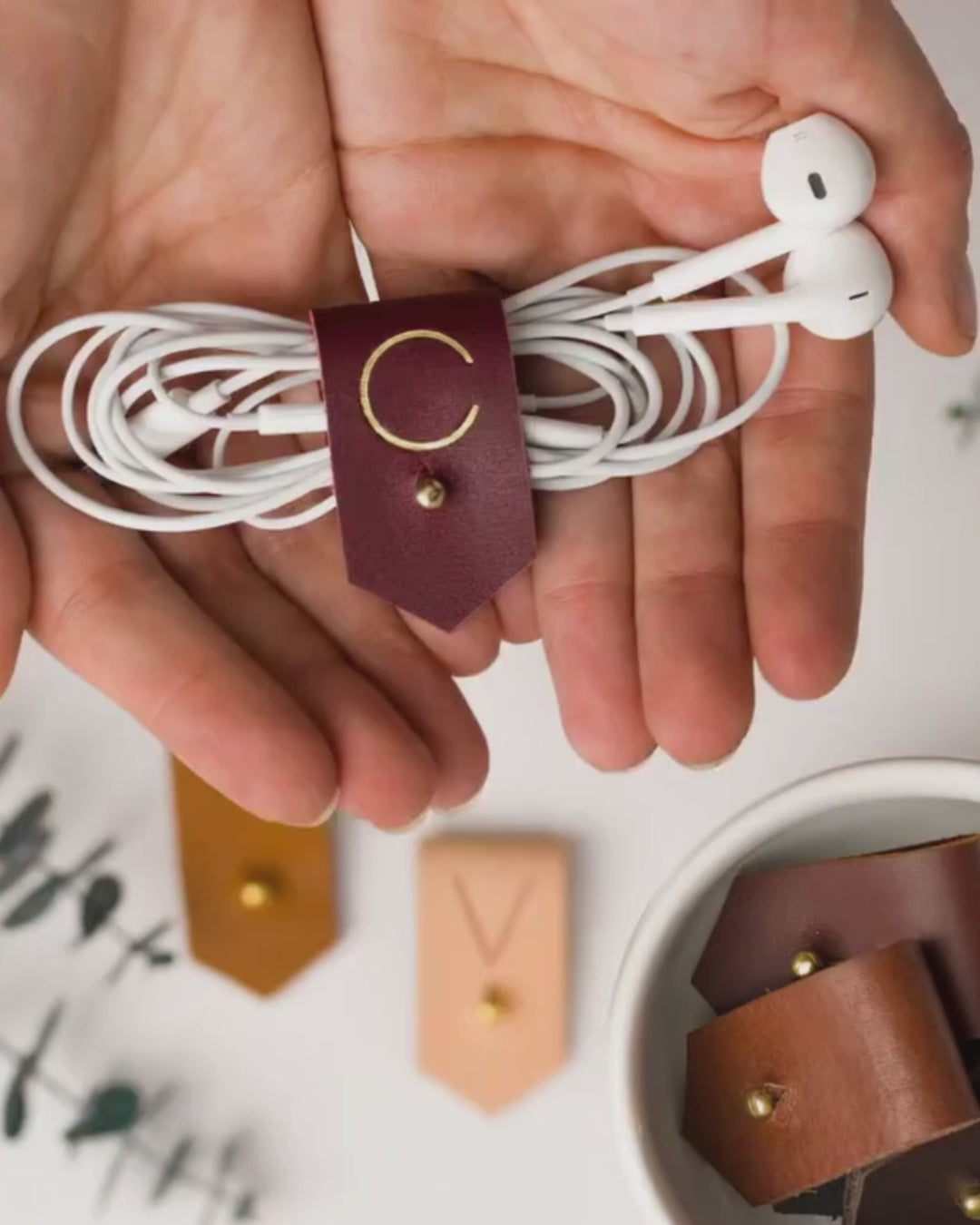 Keyaiira leather earbud holder demonstration showing a Wine C initial design and brass stud-and-post fastening for managing headphones, charger cords and everyday small cables
