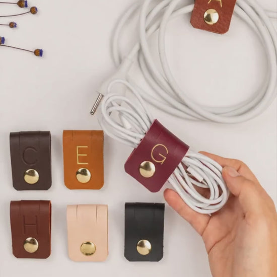 Custom Wine leather G initial cable holder shown with assorted personalized cord organizers, handmade monogram charger ties for electronics storage, desk organization and gifting by Keyaiira
