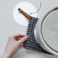Hand holding a knit potholder with a detachable leather hanging loop attached Keyaiira handcrafted leather loop accessory.