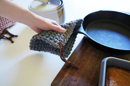 Neutral-toned woven potholder lifting cast iron pan from stovetop — heat-protective kitchen hot pad with leather hanging cord.